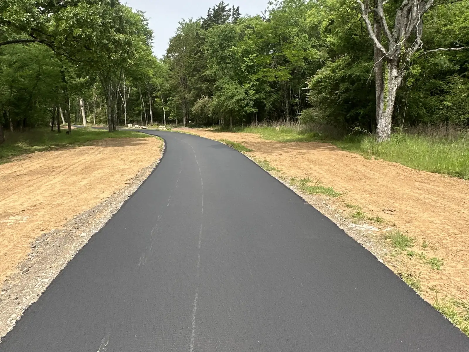 Asphalt paver laying a new asphalt driveway through the woods with a crew member walking alongside, asphalt driveway paving