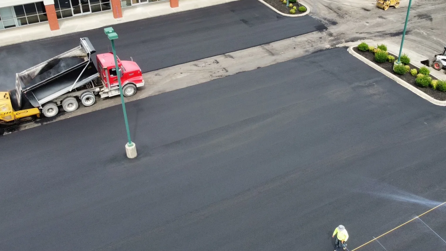 Paving crew laying hot mix asphalt across a commercial parking lot with a paver, dump truck and skid steer seen from above, asphalt paving